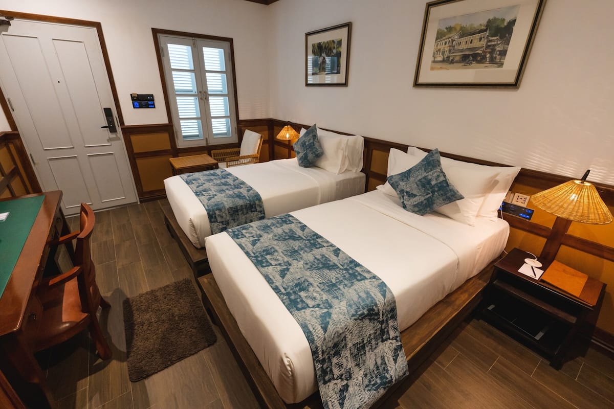 Een hotelkamer met twee bedden, een bureau en een stoel. Er is een raam met zonwering, een deur en een bedieningspaneel aan de muur.
