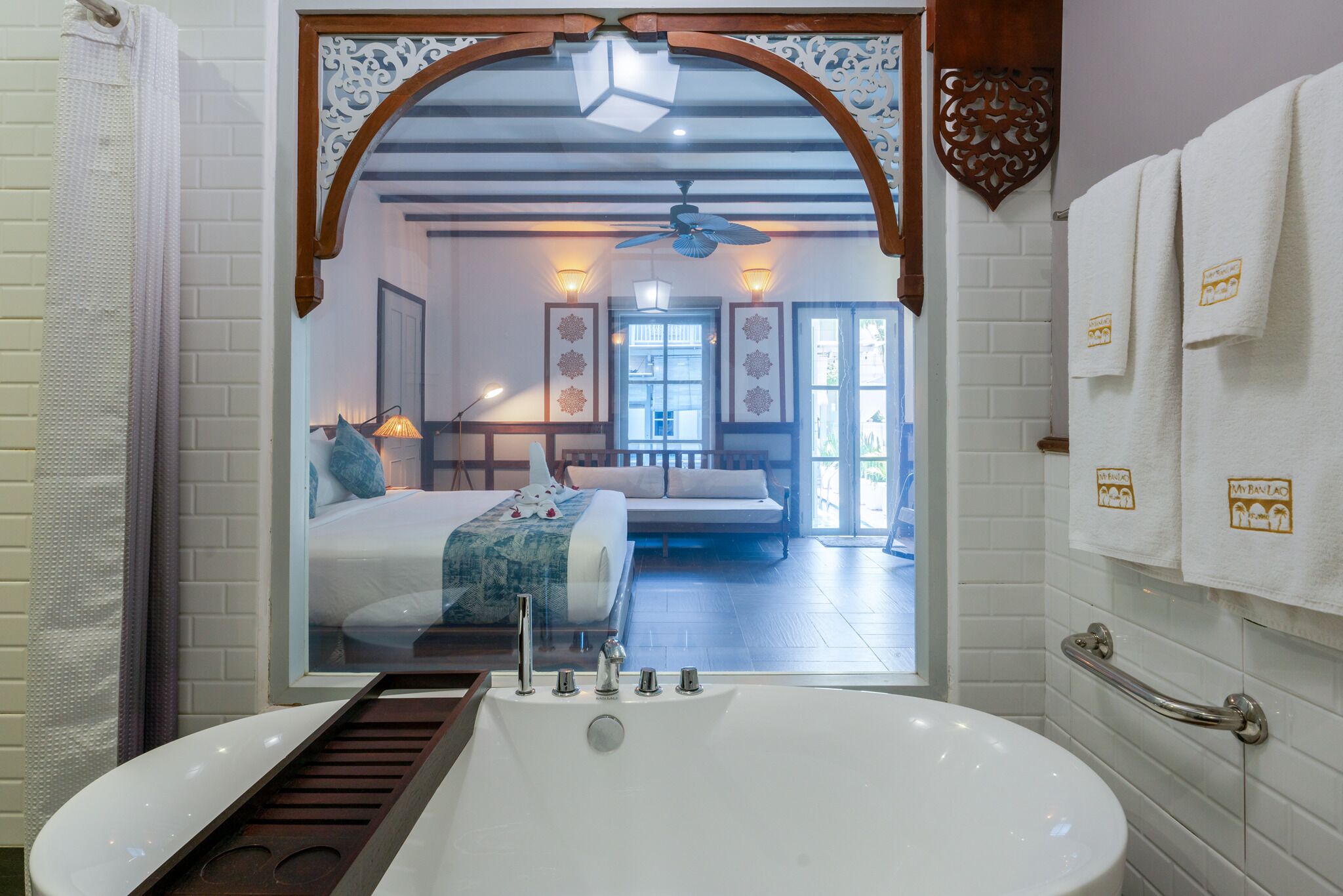 Ein modernes Badezimmer mit einer großen Badewanne, einer Dusche und einem von Glas umgebenen Spiegel, der ein Schlafzimmer mit einem Bett, einer Couch und einem Fenster reflektiert.
