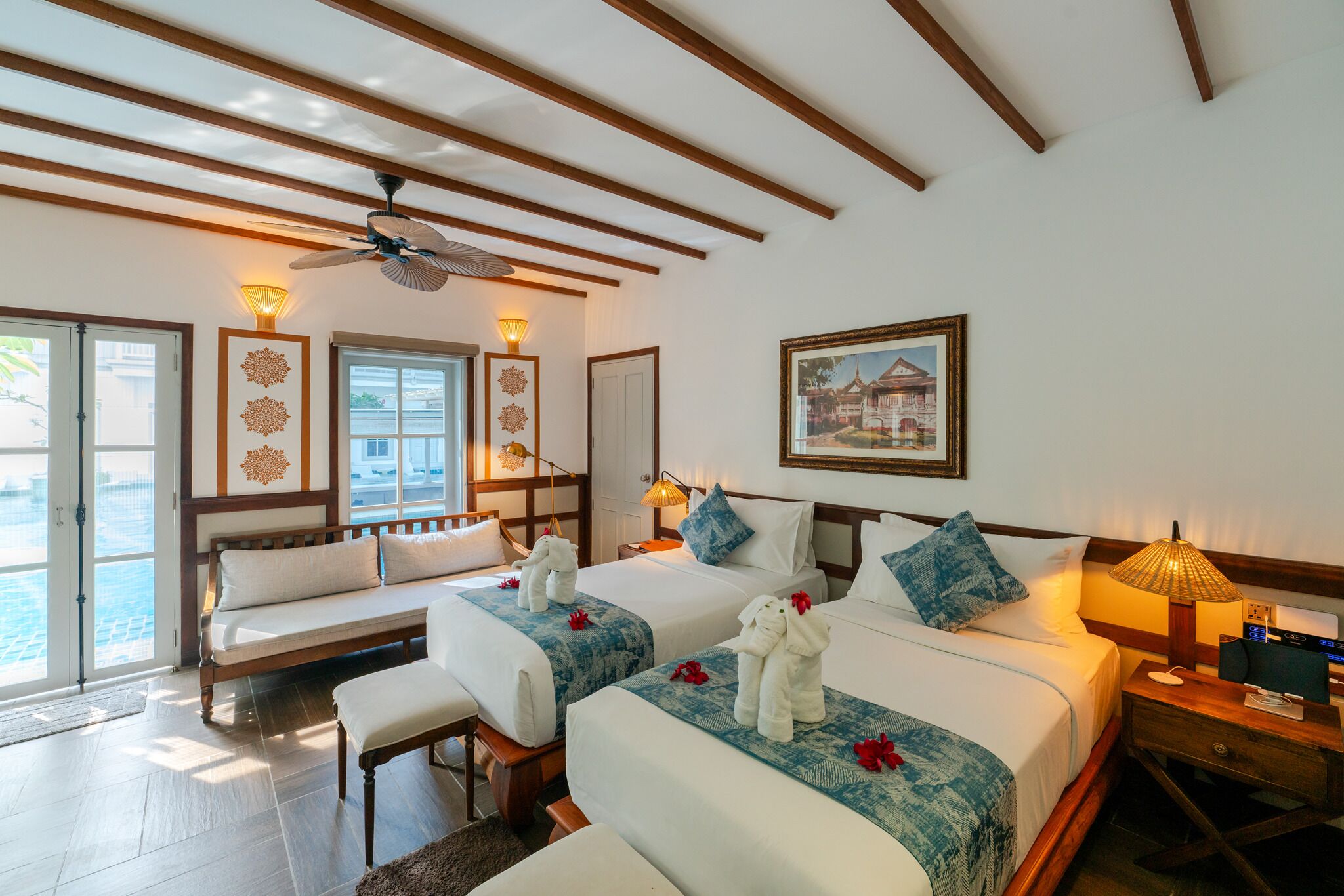 A hotel room with two beds, wooden beams, a ceiling fan, bedside lamps, a framed picture, and a bench by the window.