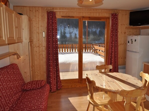A wooden cabin interior with a dining table, chairs, a sofa, a television, and a view of a snowy landscape through a large window.