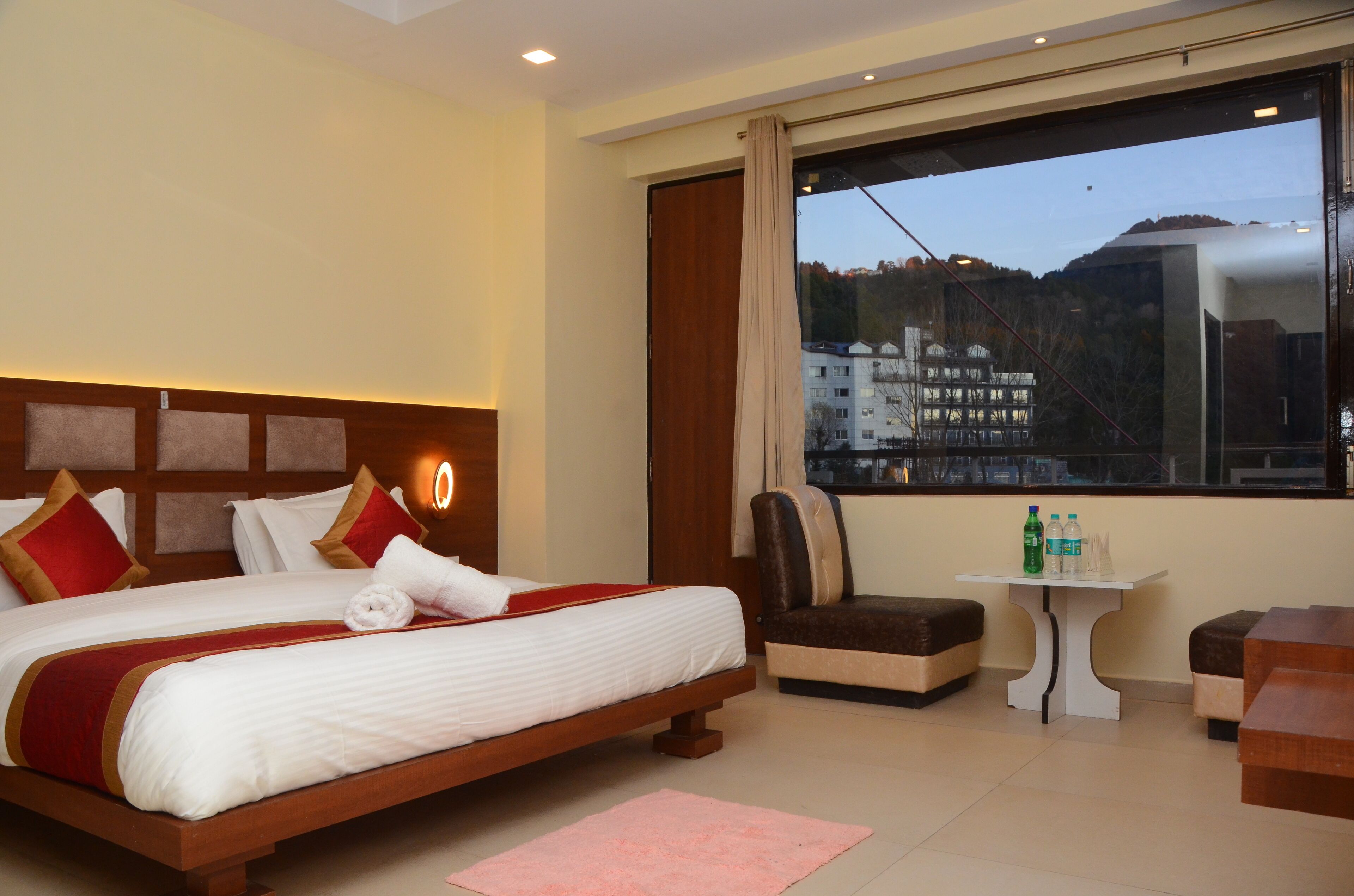 A hotel room with a large bed, a small table with bottles, and a view of buildings through a window.