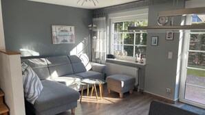 A modern living room with a grey sofa, a glass coffee table, and a small ottoman. There is a window with curtains, a radiator, and a clock on the wall.