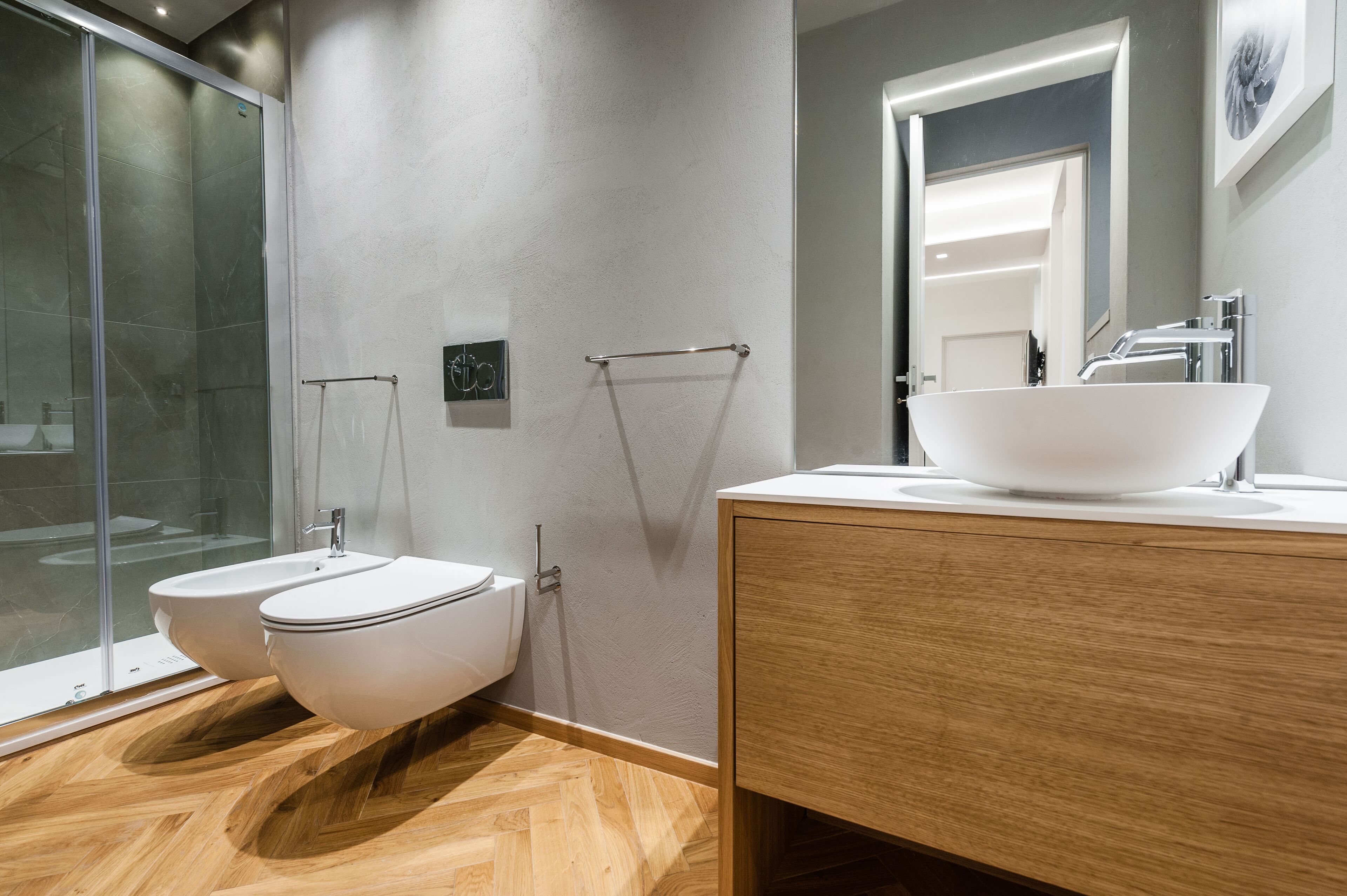 A modern bathroom with a glass shower enclosure, two white toilets, and a wooden vanity with a large sink.