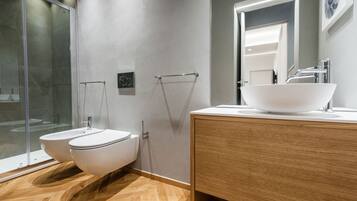 A modern bathroom with a glass shower enclosure, two white toilets, and a wooden vanity with a large sink.
