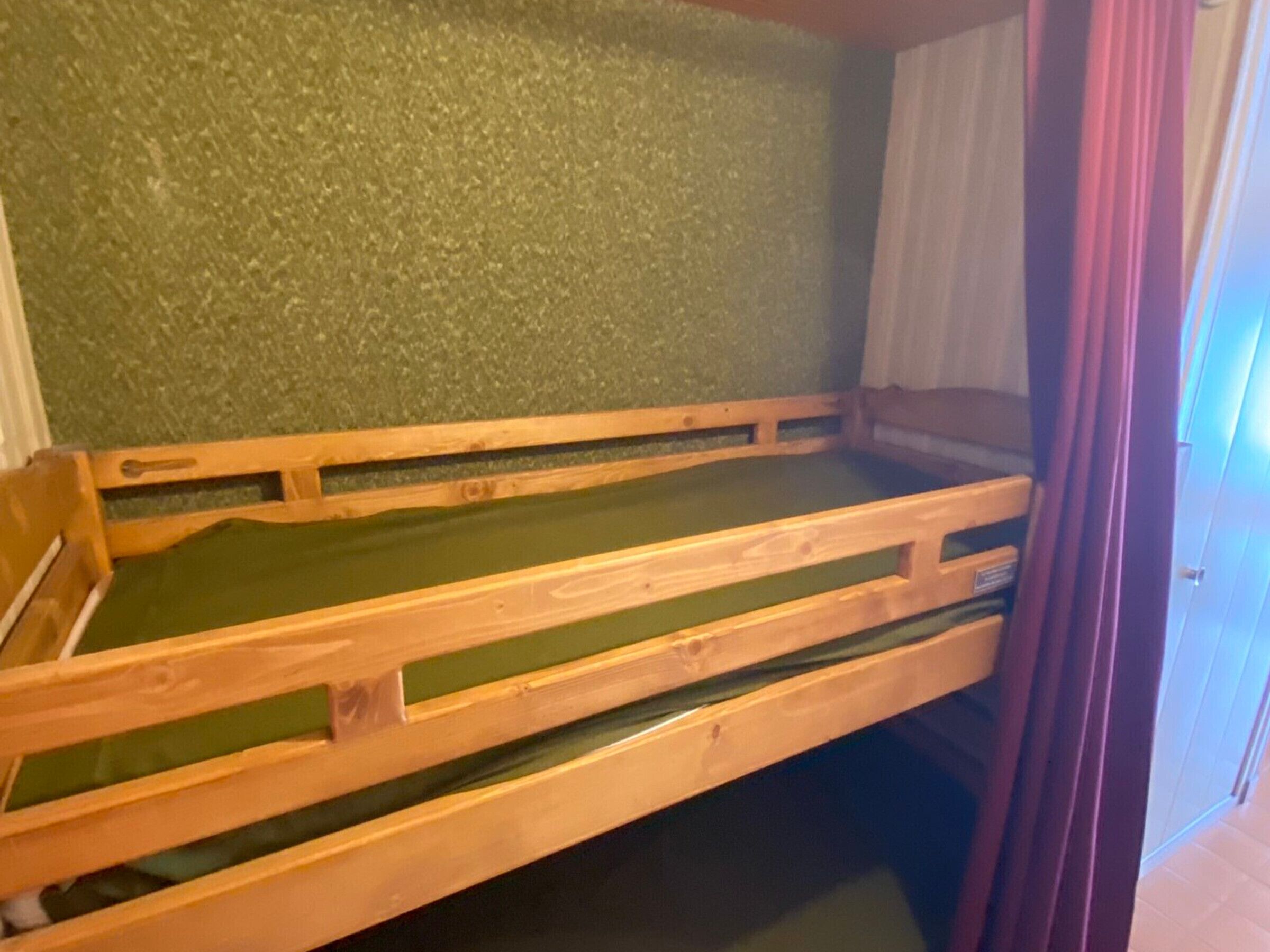 Ein Stockbett mit grüner Bettwäsche in einem Zimmer mit strukturierter grüner Wand und einer Tür mit violettem Vorhang.