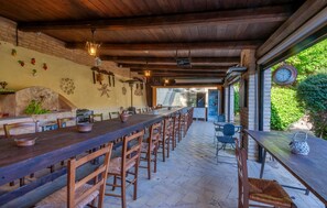 Un espace de restauration extérieur rustique, avec une longue table en bois, plusieurs chaises et une cheminée en pierre.