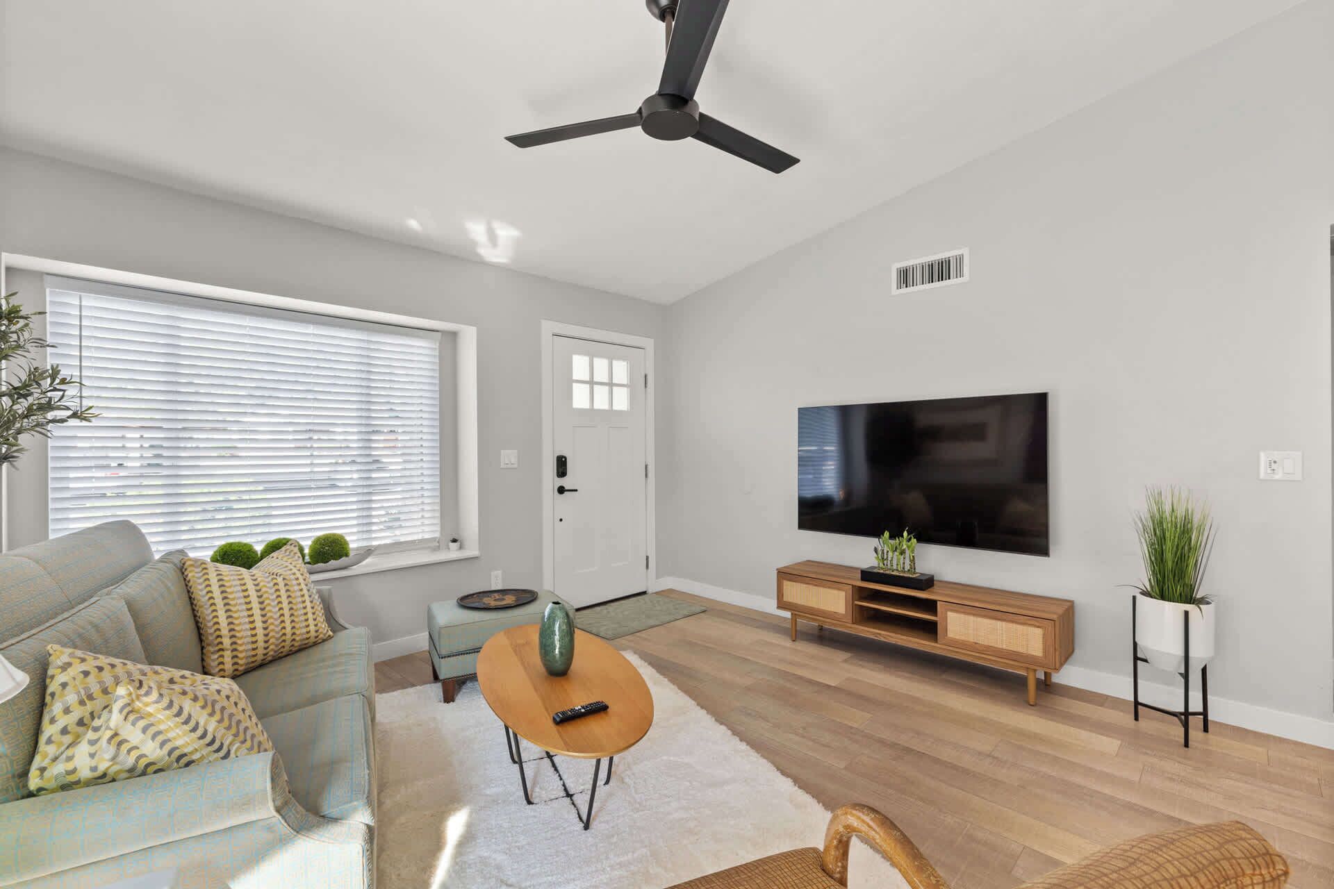 Un salon moderne avec un ventilateur de plafond, un grand téléviseur à écran plat, un meuble TV en bois, un canapé, une table basse et une plante en pot.