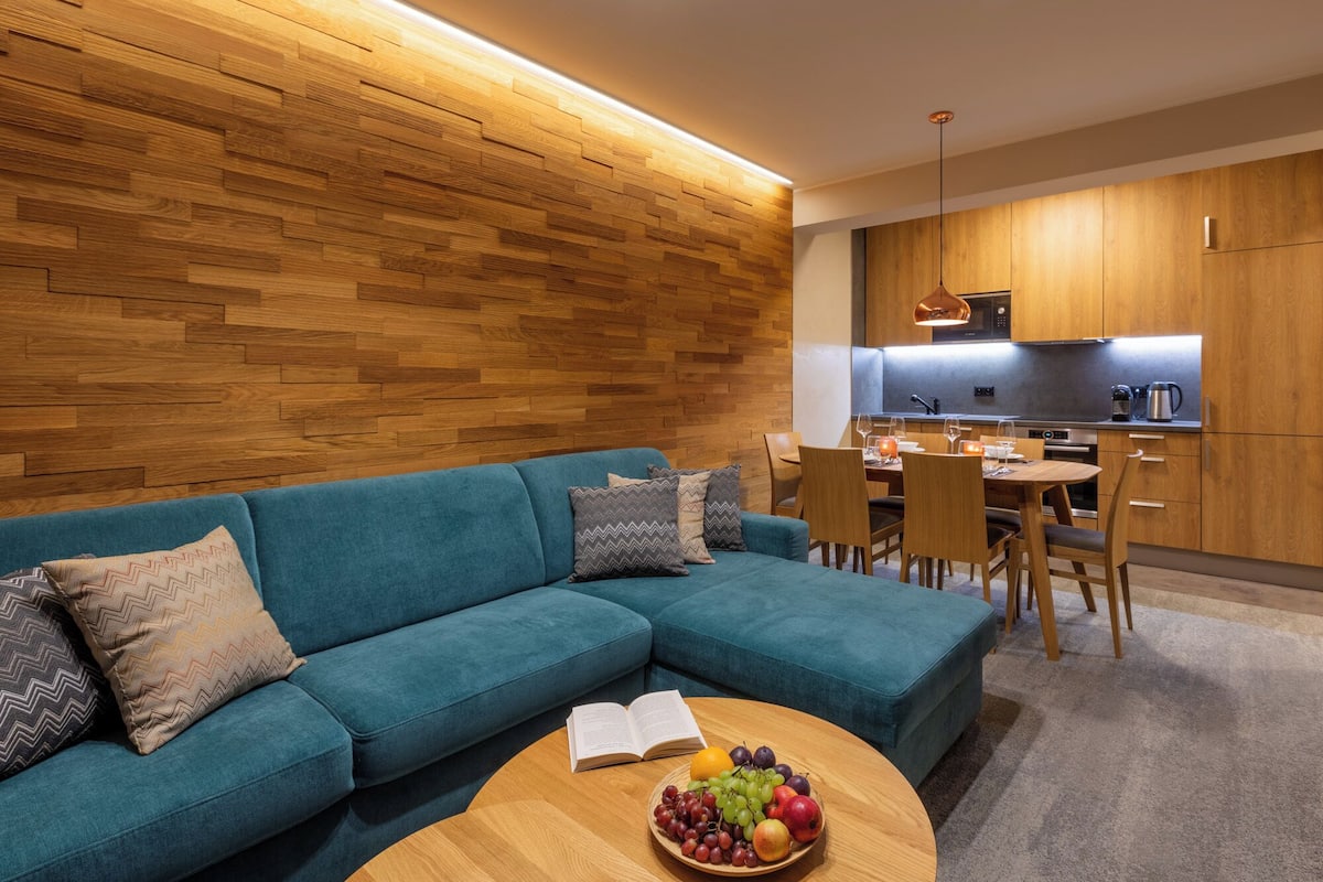 A modern living room with a teal sofa, wooden wall paneling, and a dining area with a wooden table and chairs.