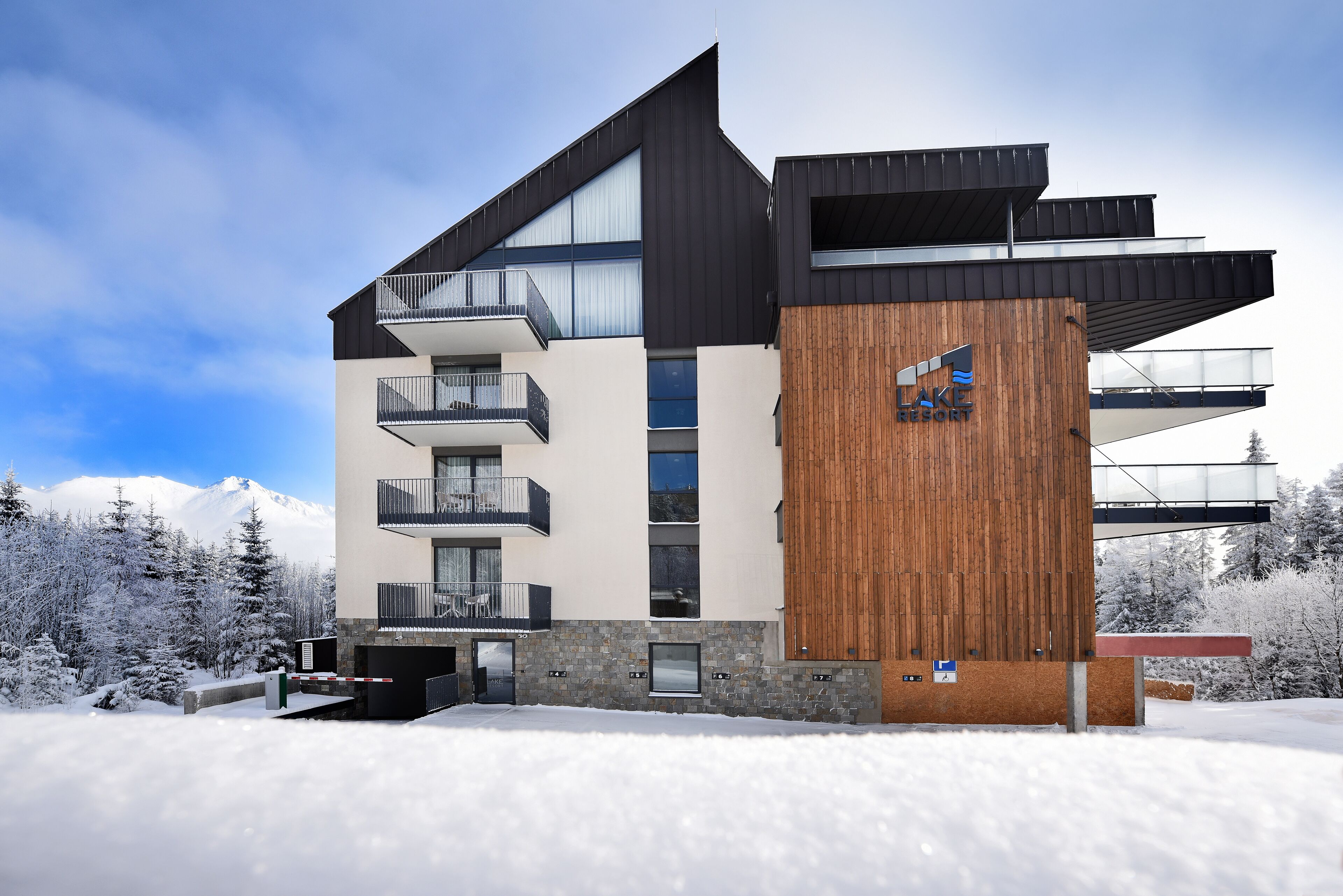 A modern multi-story building with balconies, a wooden facade, and a ski resort backdrop.