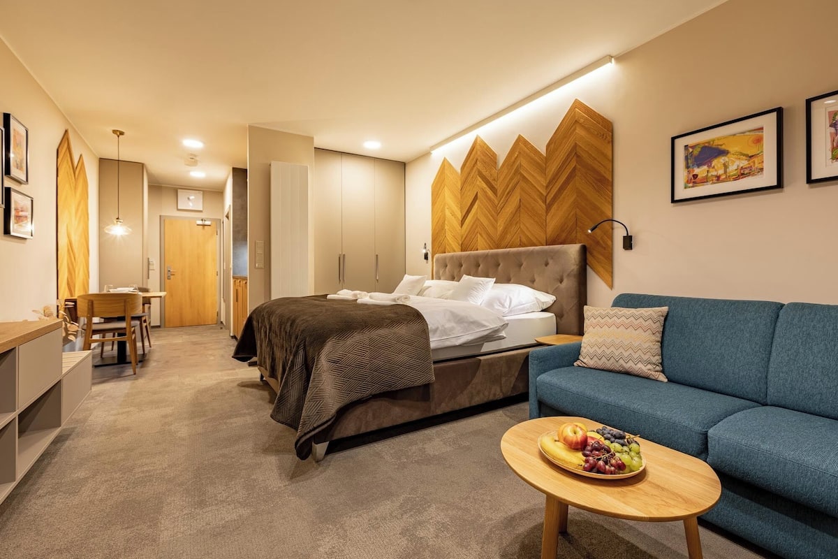 A modern hotel room with a bed, a sofa, a dining area, and a small table with fruit.