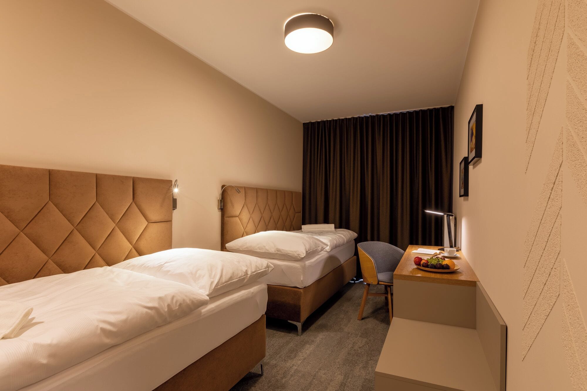 A hotel room with two beds, a desk with a chair, a lamp, and a small table with fruits.