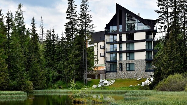 A modern multi-story building with a stone facade, large windows, and a sloped roof, surrounded by a forest and a body of water. - APLEND Lake Resort  (Štrba)