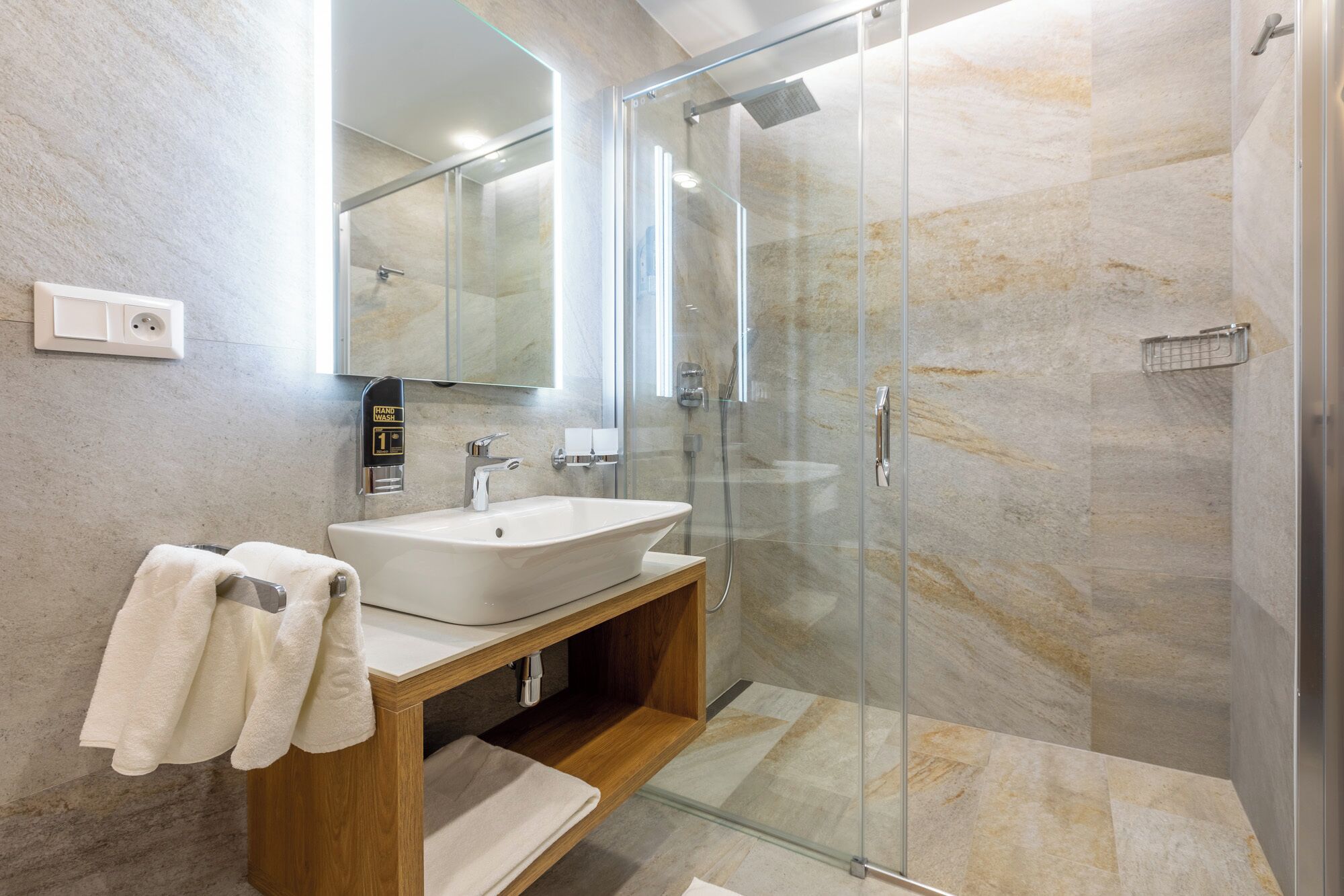 Un baño moderno con ducha envuelta en vidrio, un lavabo blanco y un mueble de tocador de madera.