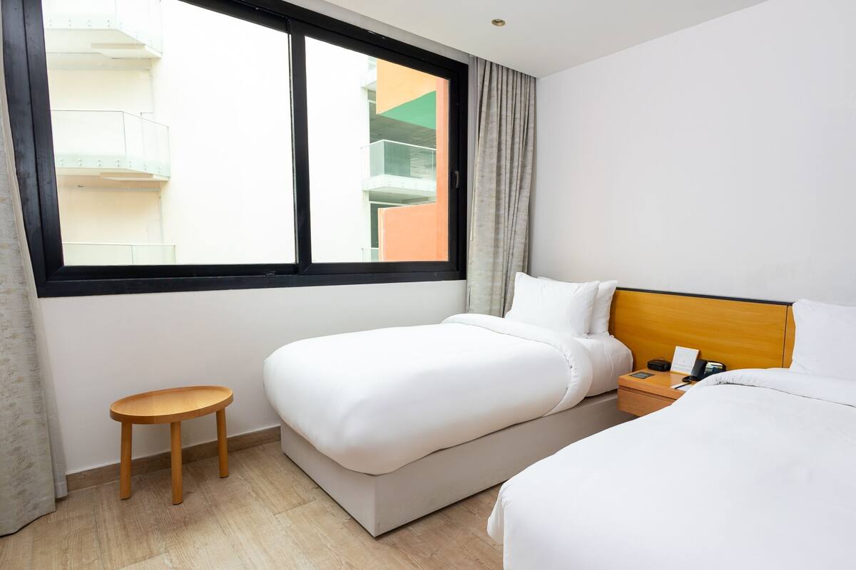 A hotel room with two beds, a small wooden stool, a nightstand with a phone and a lamp, and a window with a view of a building.
