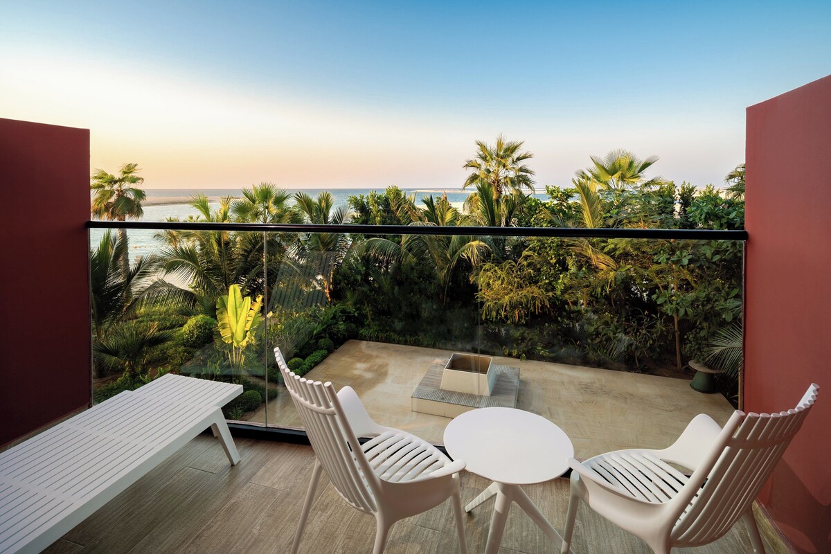 A balcony with a view of the sea, a white bench, two white chairs, and a small table.