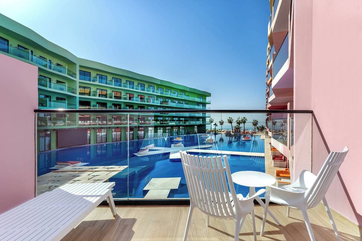 A modern hotel balcony with a pool, white lounge chairs, and a view of the pool area and hotel building.