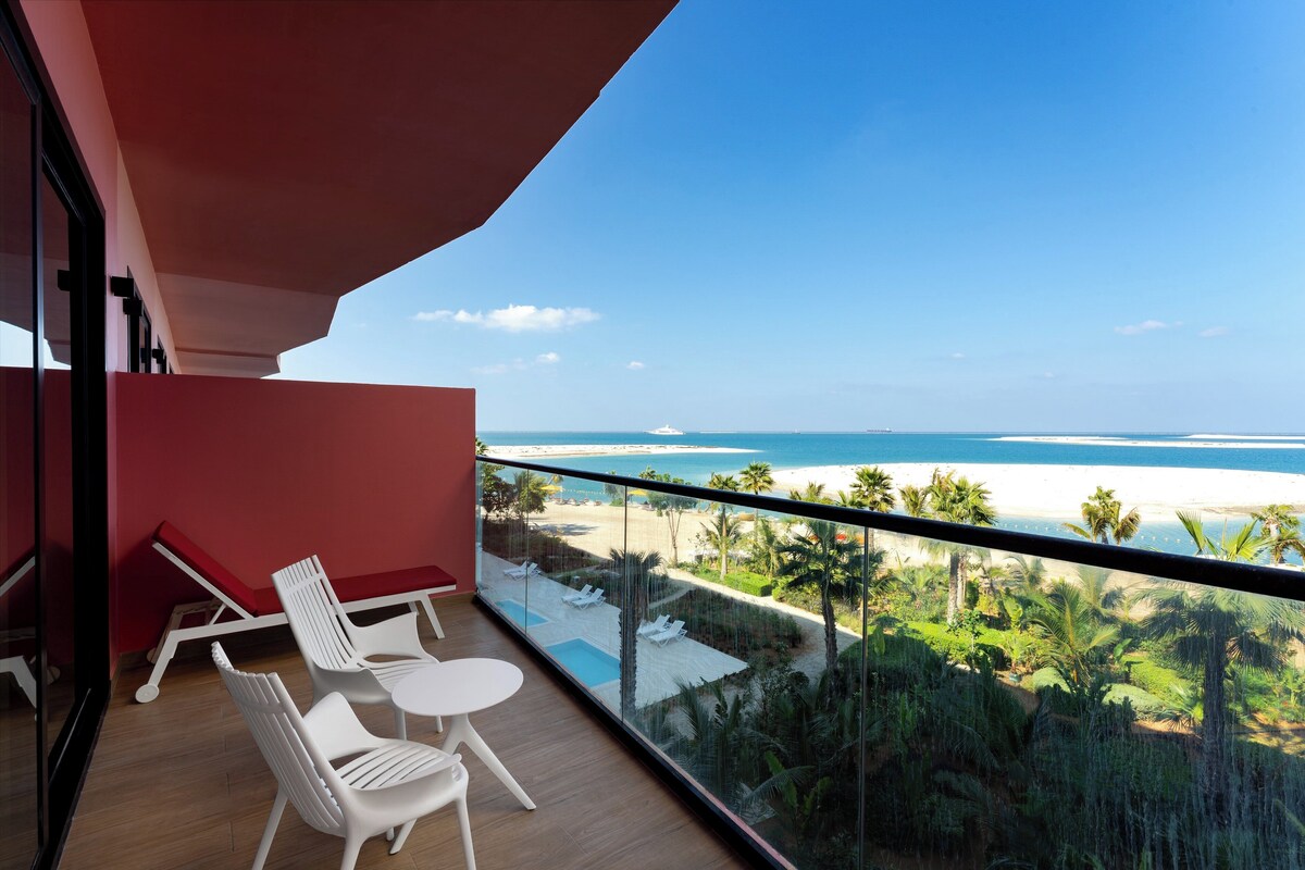 A balcony with white chairs and a table, overlooking a beach and palm trees.
