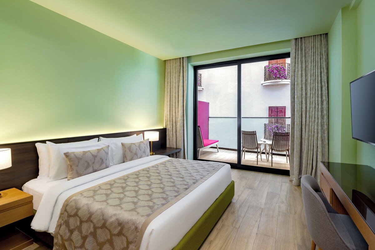 A hotel room with a large bed, a TV, a nightstand, and a balcony with a chair and table.