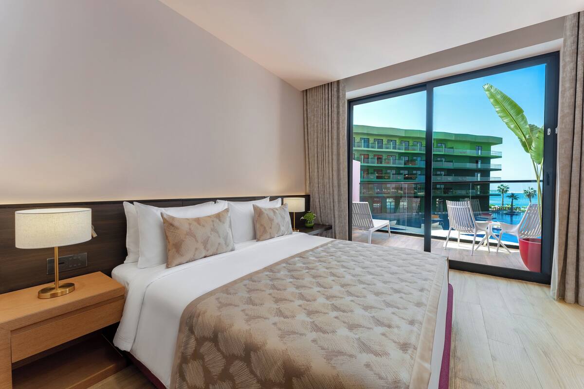 A hotel room with a large bed, a nightstand with a lamp, a view of a pool and building through a large window.