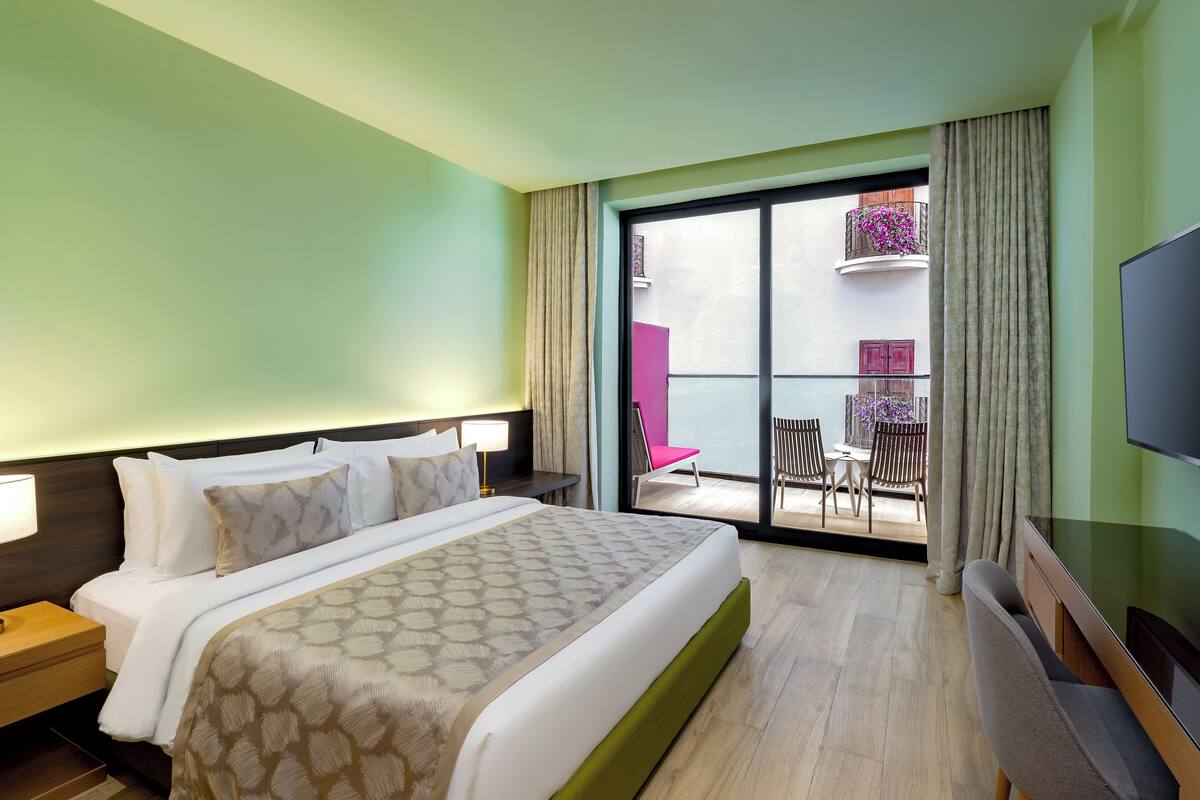 A hotel room with a large bed, a TV, a nightstand, and a balcony with a chair and table.