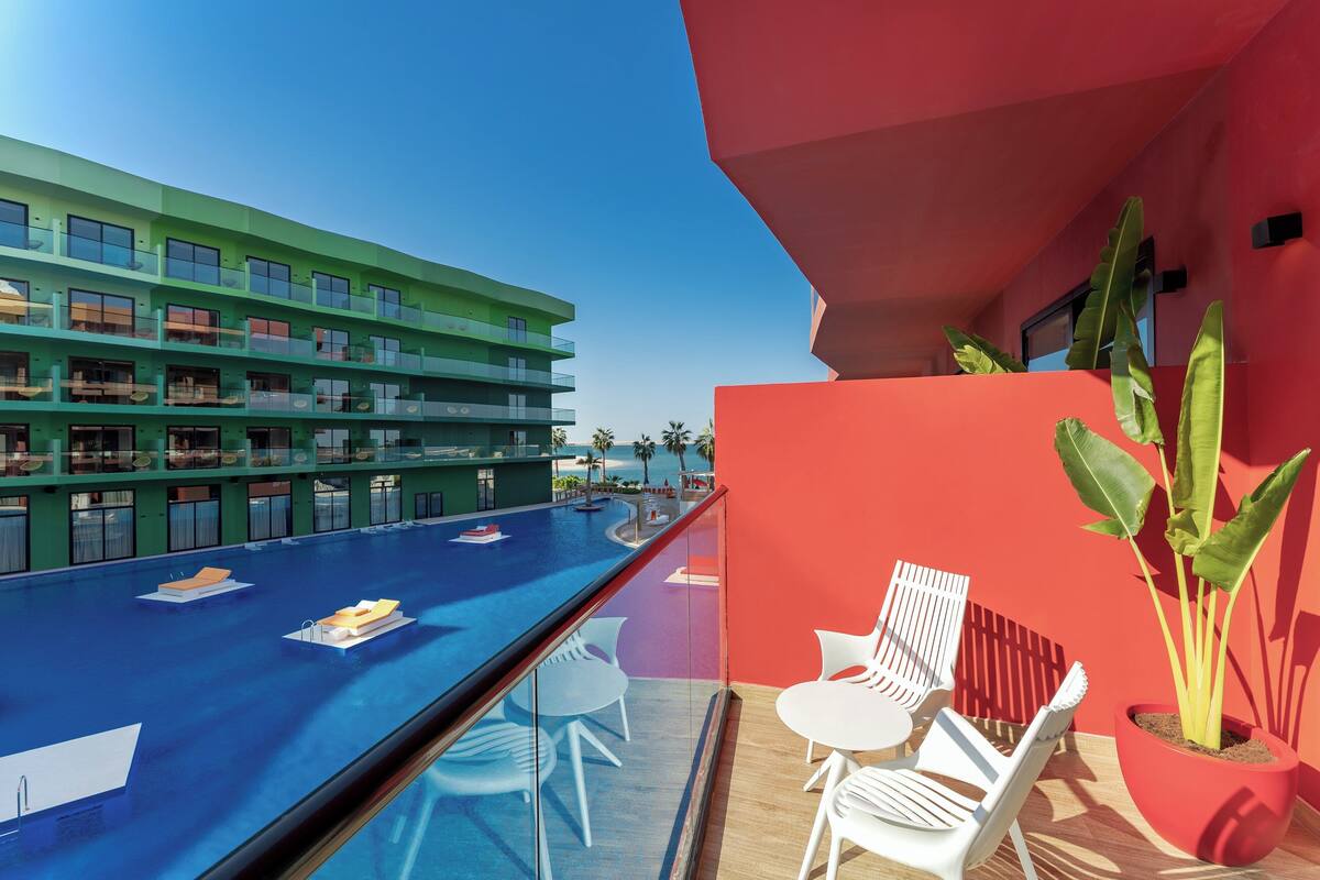 A balcony with a pool, white chairs, and a red wall with a plant.