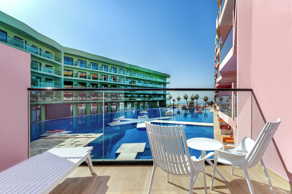 A modern hotel balcony with a pool, white lounge chairs, and a view of the pool area and hotel building.