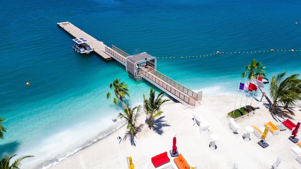 A beach resort with a pier extending into the ocean, a sandy beach with lounge chairs and umbrellas, and a small island in the background. - voco Monaco Dubai Heart of Europe by IHG (Dubai)