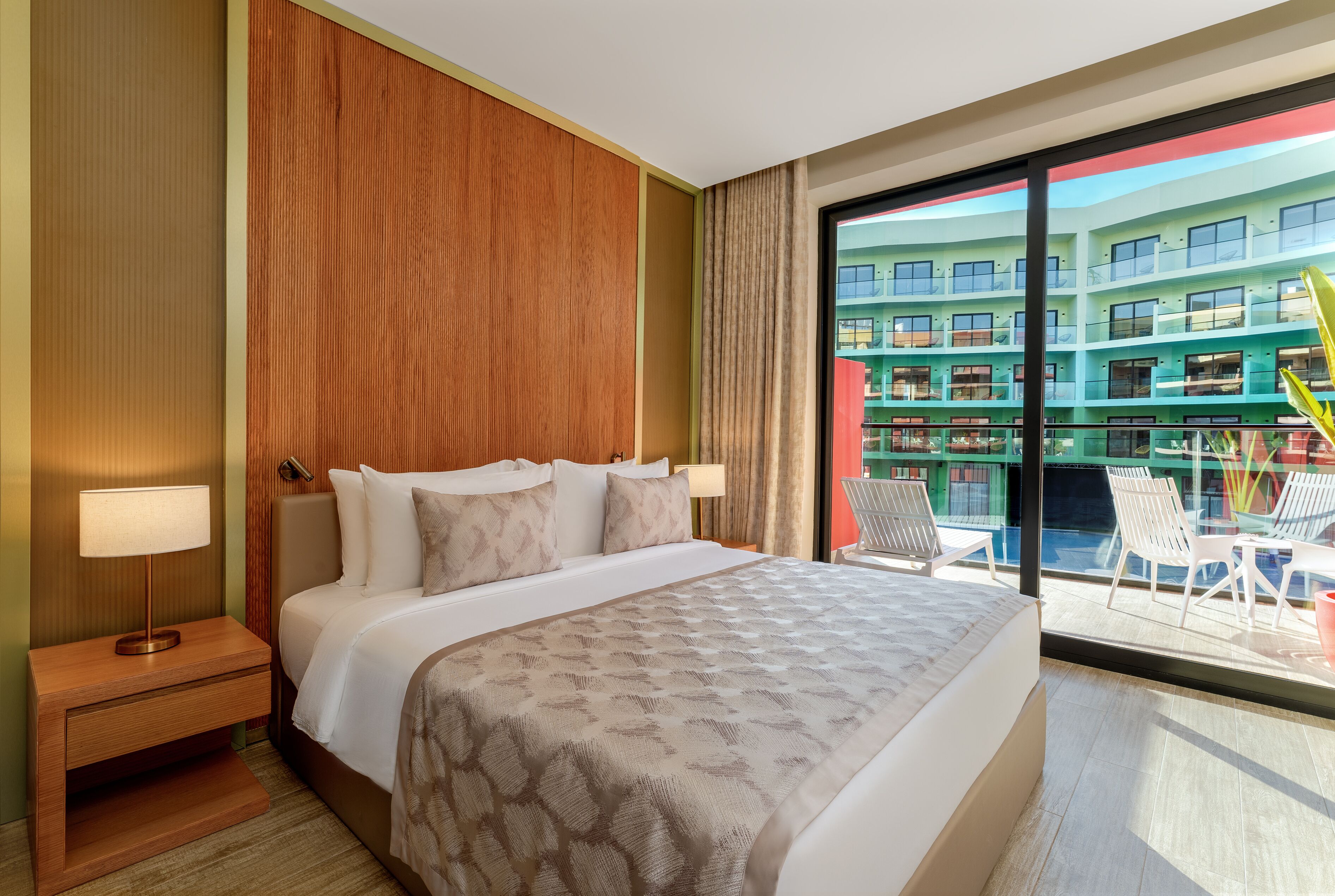 A hotel room with a large bed, a nightstand, a lamp, and a view of a building with balconies.