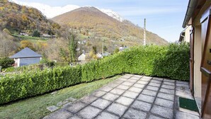 A paved patio with a view of a mountainous landscape, a hedge, and a building with a wooden door. - Cozy Chalet with 3 Bedrooms, Terrace, Parking, Near Cauterets Center (Cauterets)