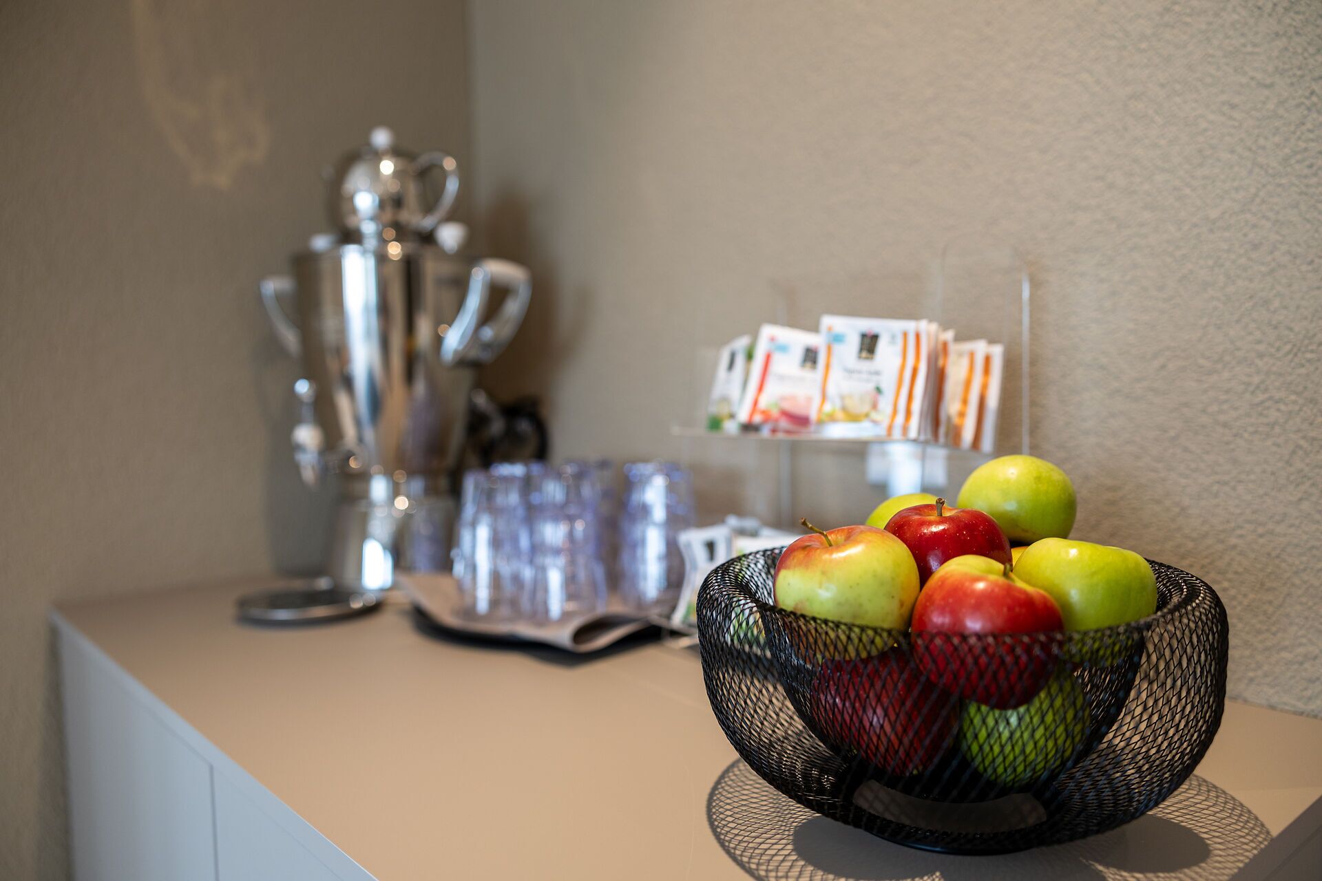 A wire basket filled with apples on a counter, with a coffee machine and magazines in the background.