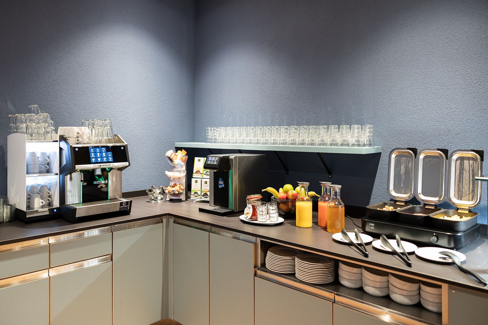 Un espace moderne dédié au petit-déjeuner, avec une machine à café, un présentoir de fruits et un distributeur de jus.