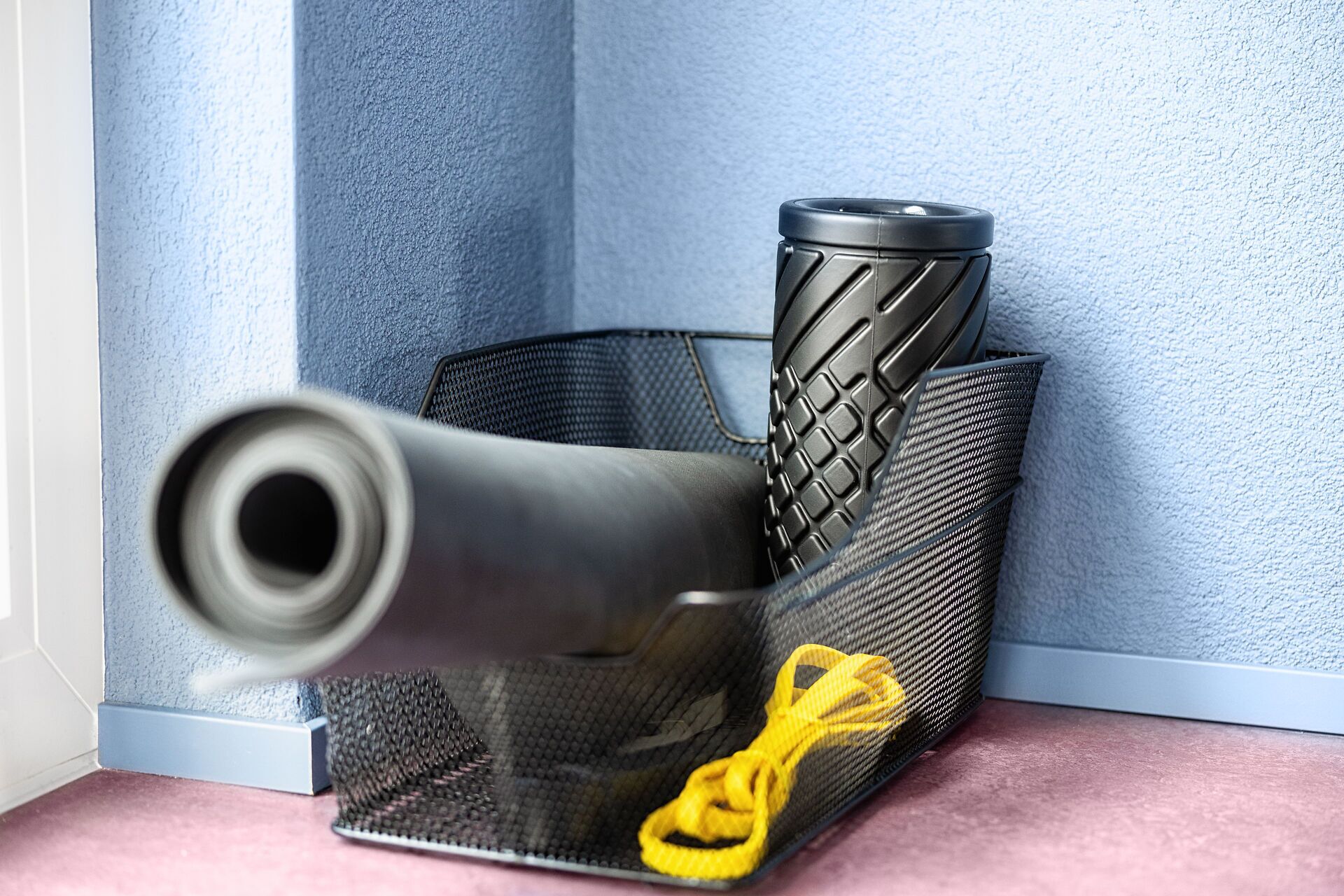 Een hoek van een kamer met een blauwe, gestructureerde muur, een zwarte netmand met een geel object, en een cilindrische container met een reliëfpatroon.