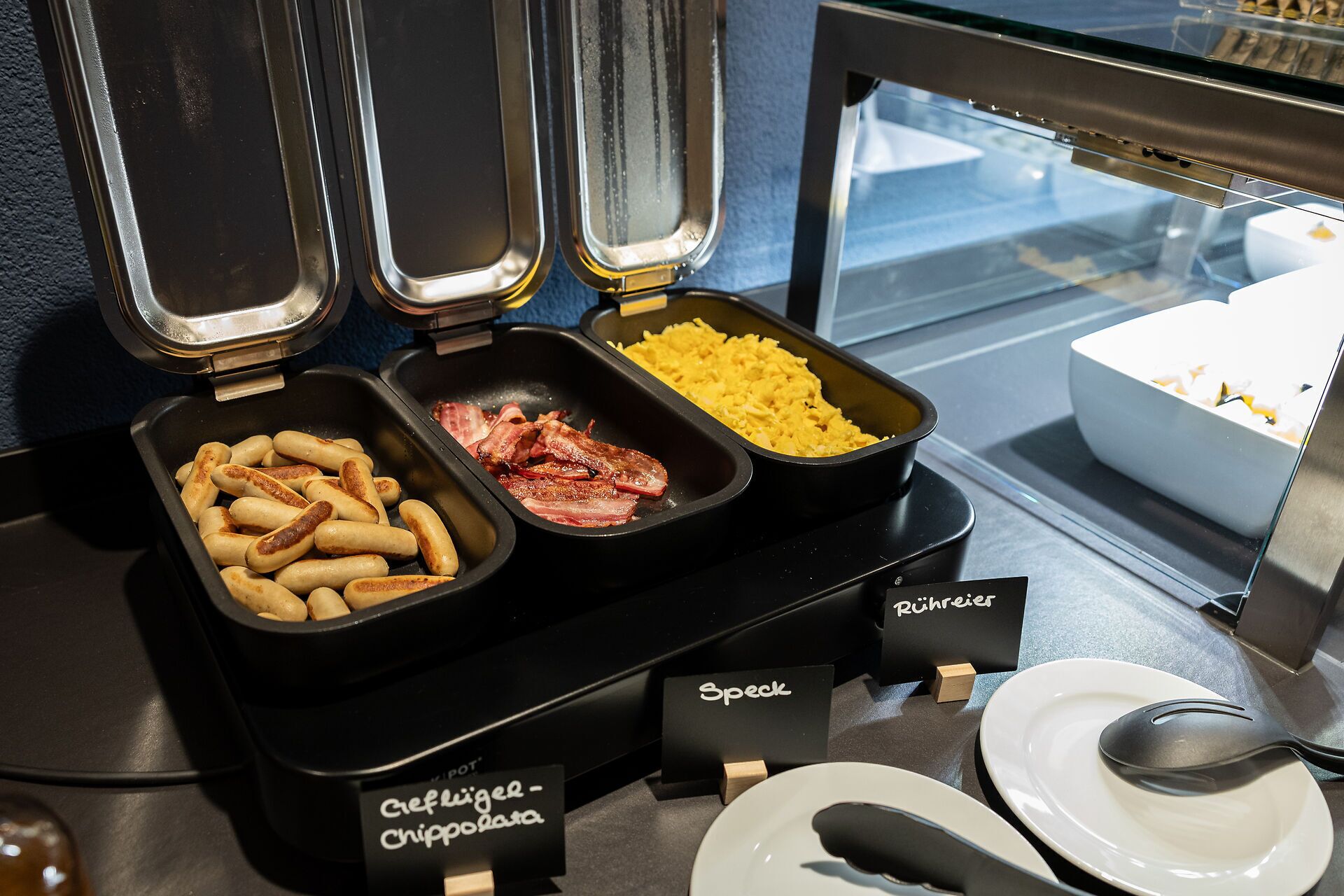 A buffet with three trays of food: sausages, bacon, and scrambled eggs.