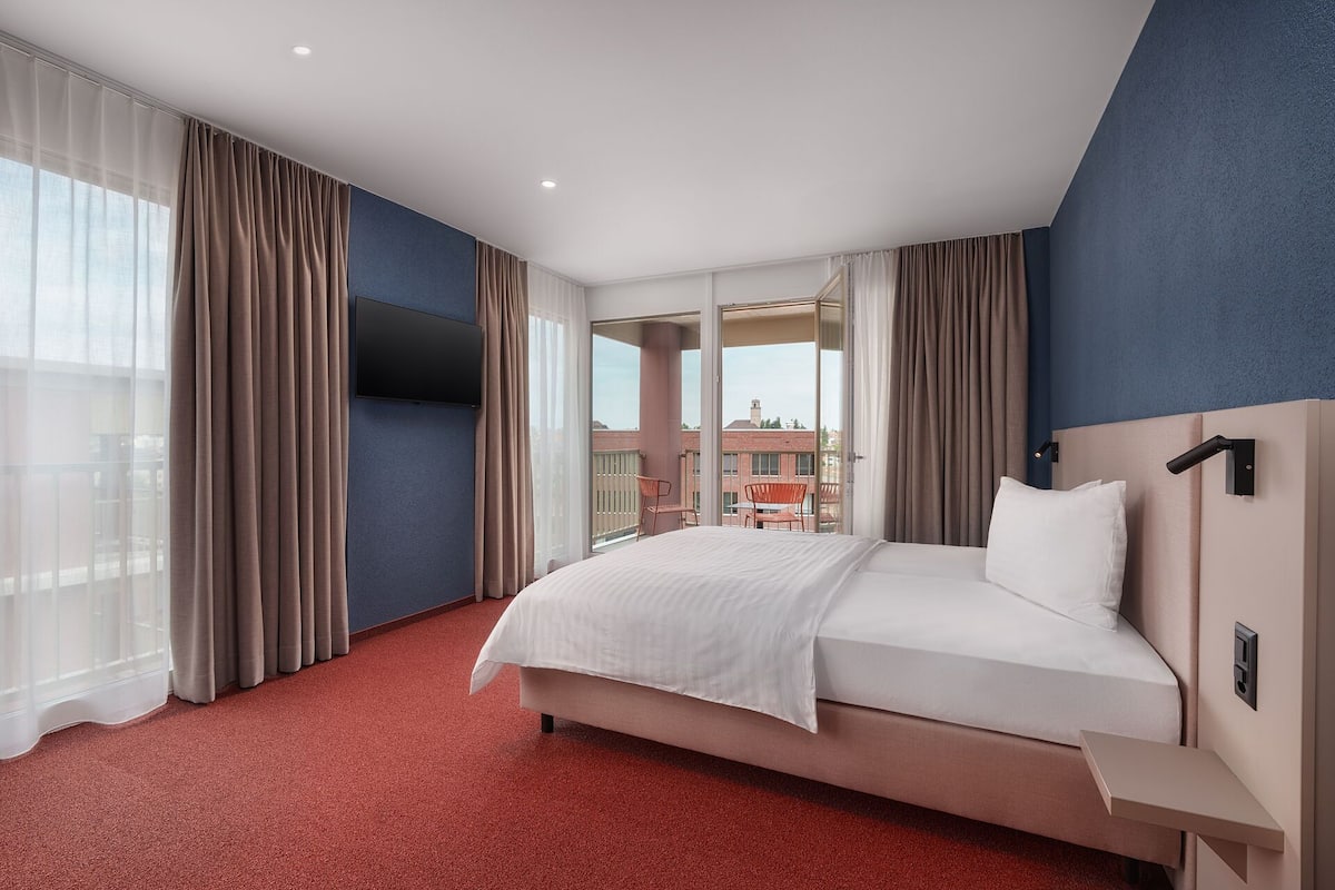 Habitación de hotel con una cama grande, un televisor montado en la pared, un balcón con sillas y una alfombra roja.
