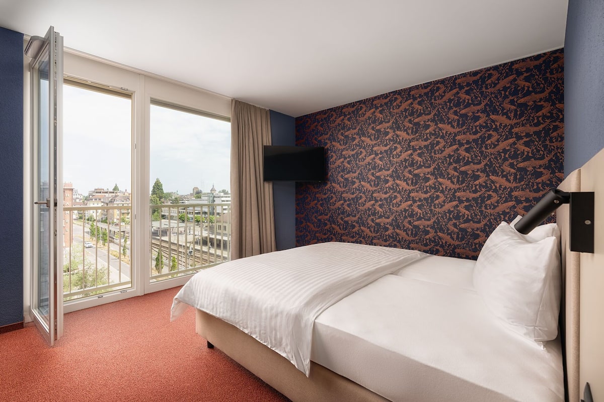 Habitación de hotel con cama, televisor montado en la pared y balcón con vista a la ciudad.