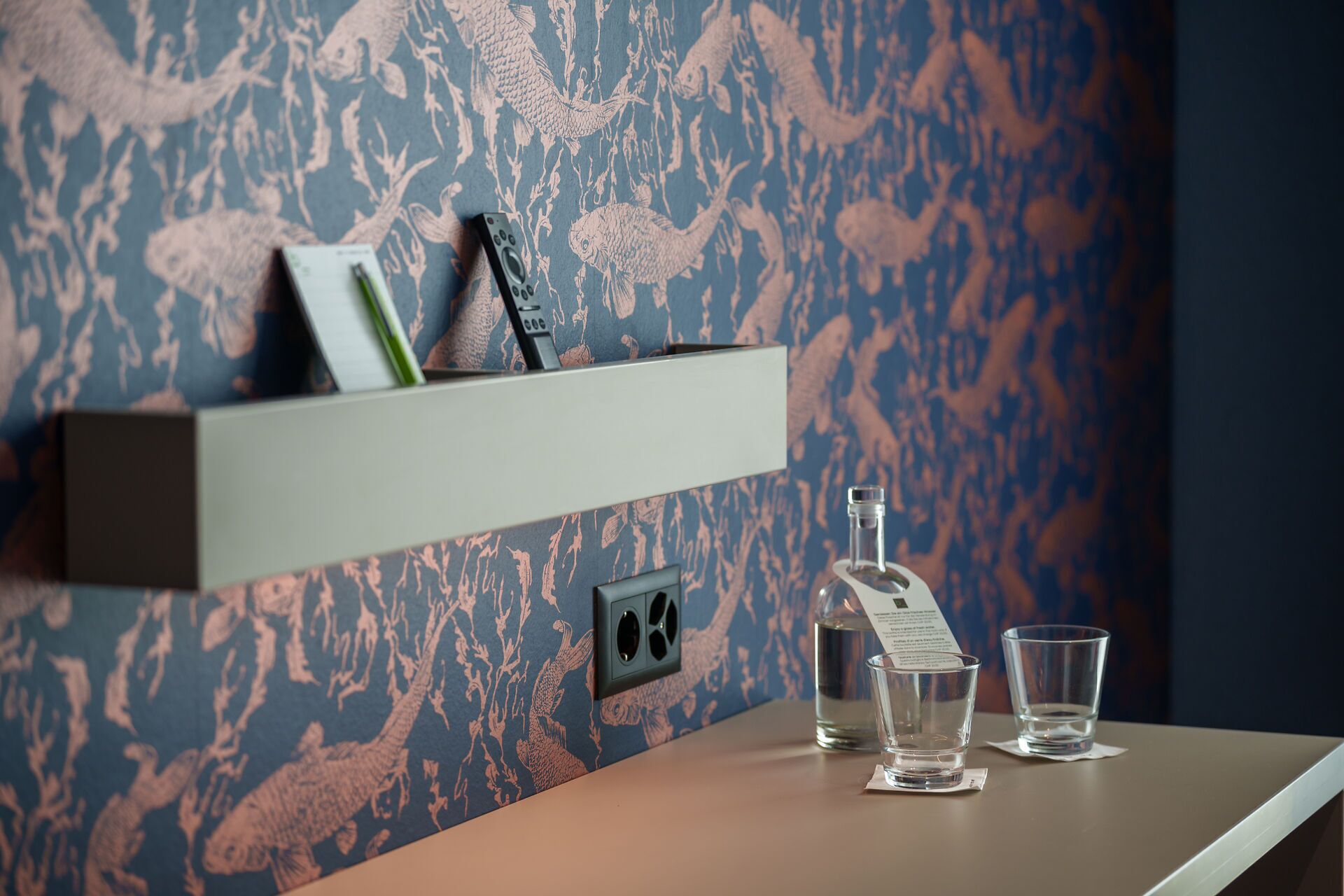 Una pared con un estampado de peces azules y rosados, un estante con un bolígrafo, un control remoto y dos vasos sobre una mesa.