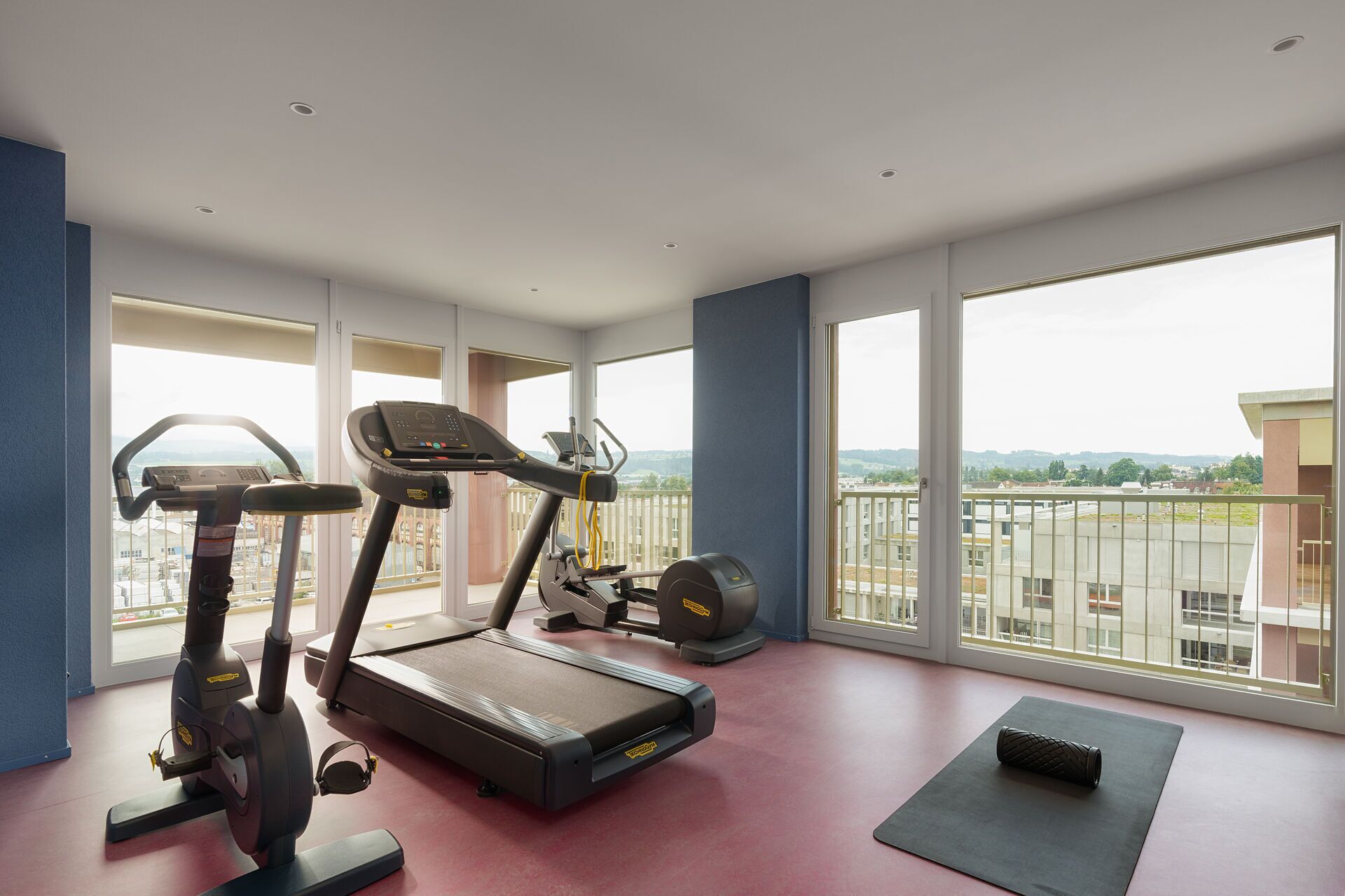 A spacious gym with a treadmill, elliptical, and stationary bike, large windows with a city view.
