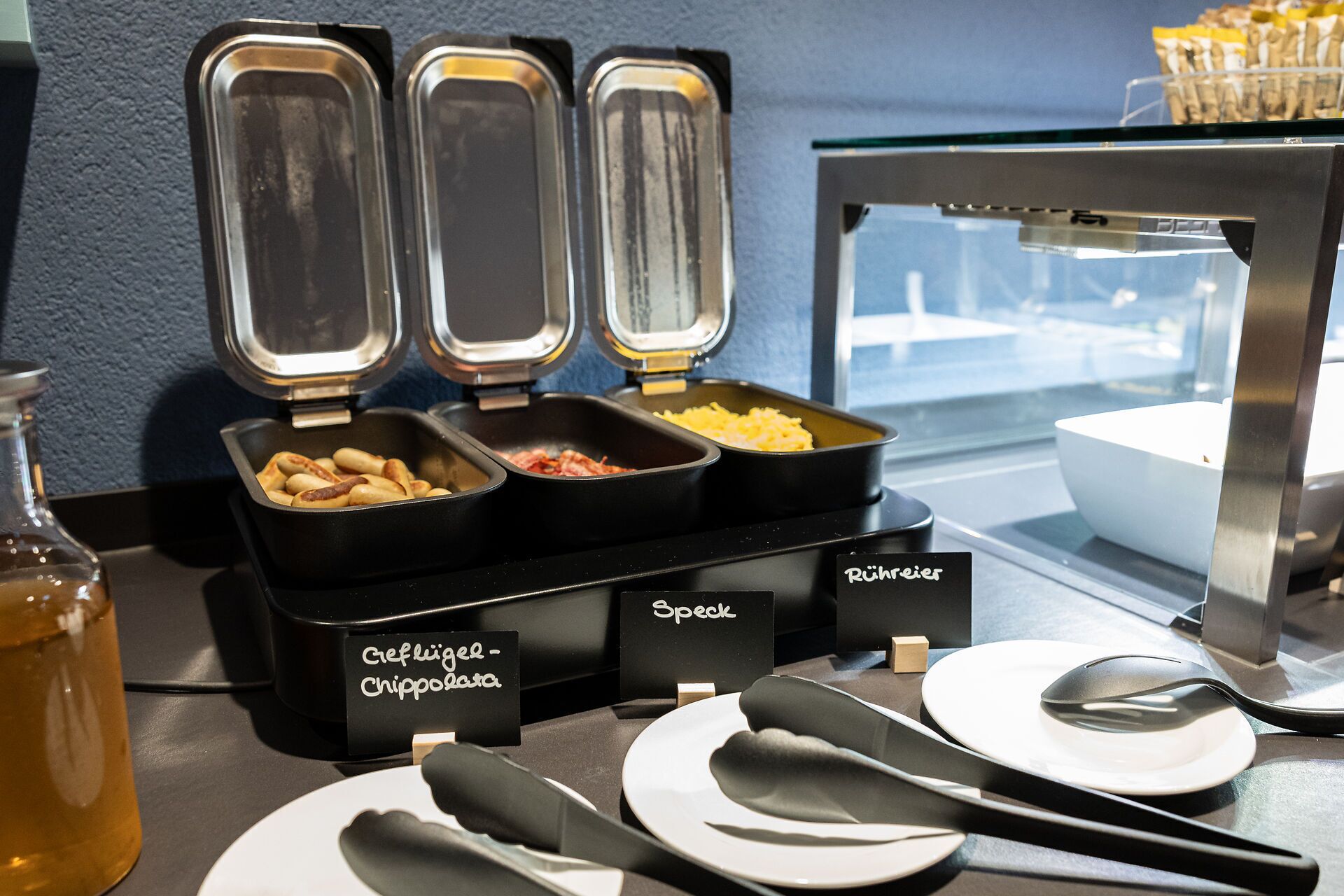 Buffetopstelling met containers voorzien van labels voor 'Geflügel - Chipollara' en 'Rühreies Speck'.