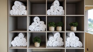 A hotel room with a beige wall and a large window. There are multiple shelves with neatly rolled white towels and small potted plants. - bsmart hotel Arbon (Arbon)