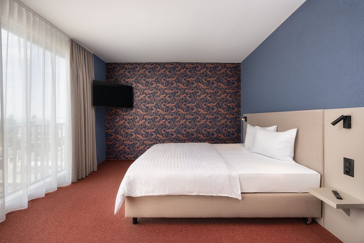 Una habitación de hotel con una cama, un televisor montado en la pared y un ventanal con cortinas.