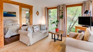 Un salon confortable avec un canapé, une table basse et un téléviseur. On aperçoit un lit au fond et une fenêtre donnant sur des arbres.