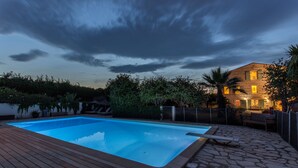 Une piscine avec une terrasse en bois, entourée d’un patio en pierre et d’une maison dont les fenêtres sont illuminées.