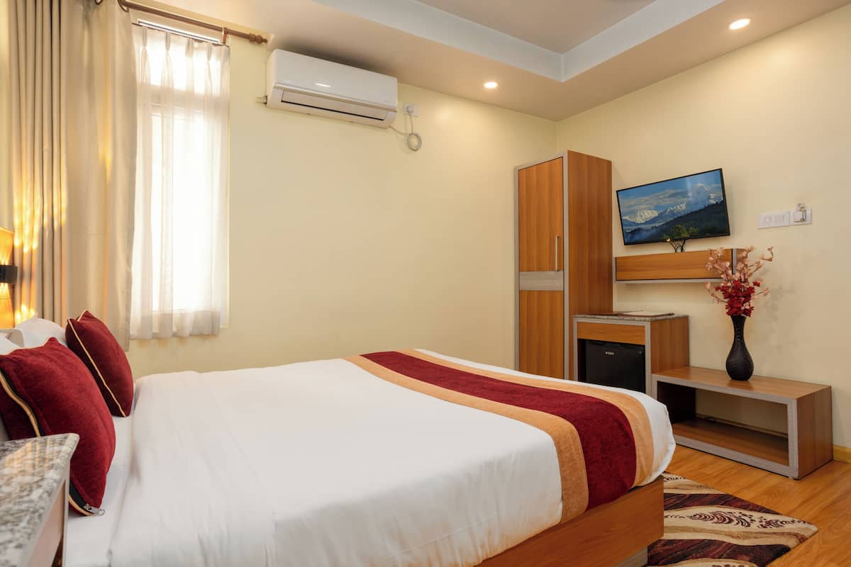 A hotel room with a bed, a television, an air conditioning unit, a wooden wardrobe, and a small desk with a vase of flowers.