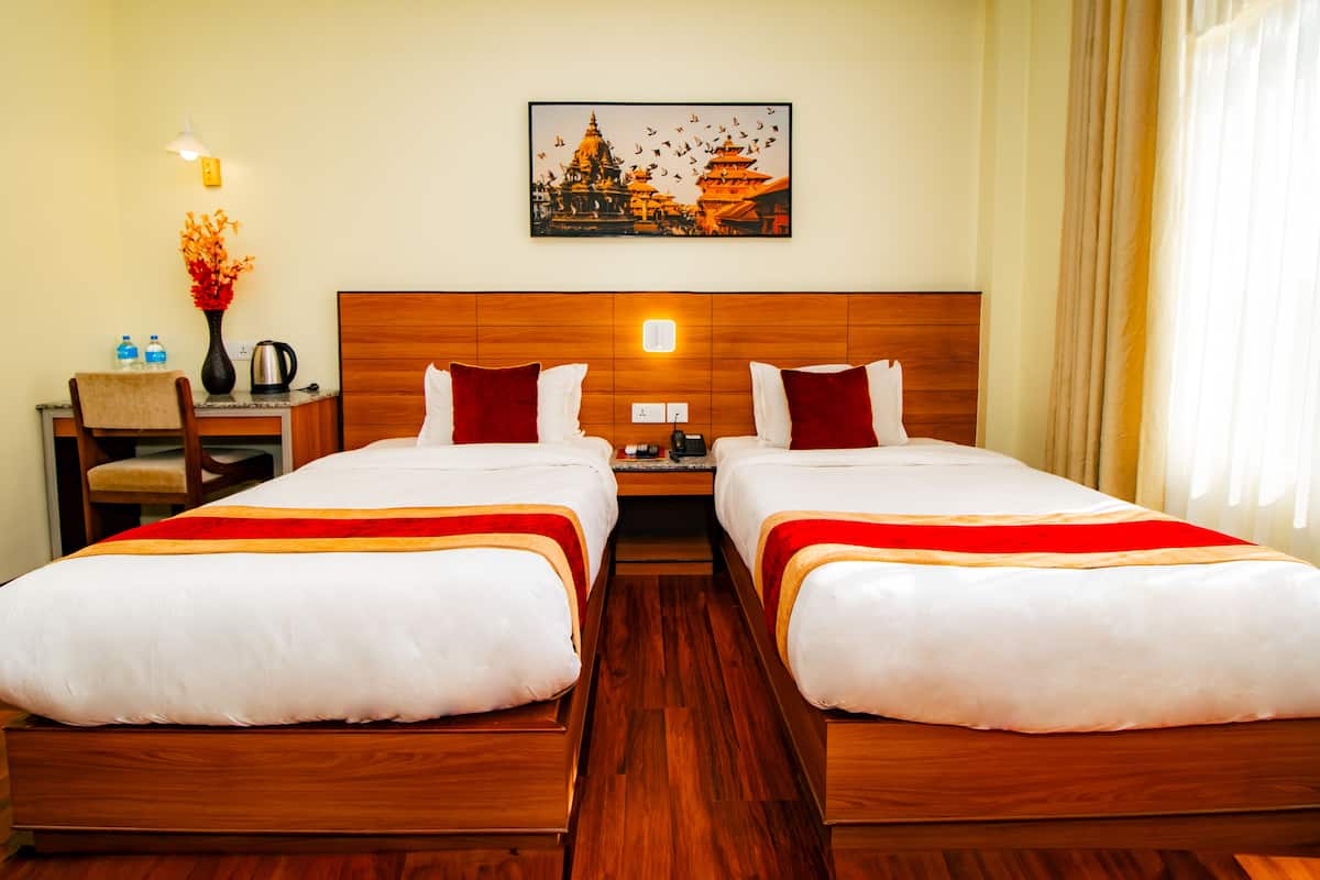 A hotel room with two beds, a wooden headboard, a nightstand, a desk, a chair, a vase with flowers, a painting of a temple, and a window with curtains.