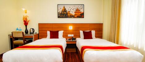 A hotel room with two beds, a wooden headboard, a nightstand, a desk, a chair, a vase with flowers, a painting of a temple, and a window with curtains.