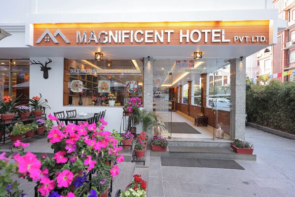 The exterior of the Magnificent Hotel Pvt. Ltd. with a visible entrance, outdoor seating area, and potted plants.
