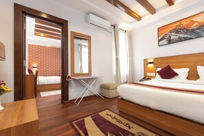 A hotel room with a wooden bed, a small desk, an ironing board, a brick wall, and a painting of a mountain.