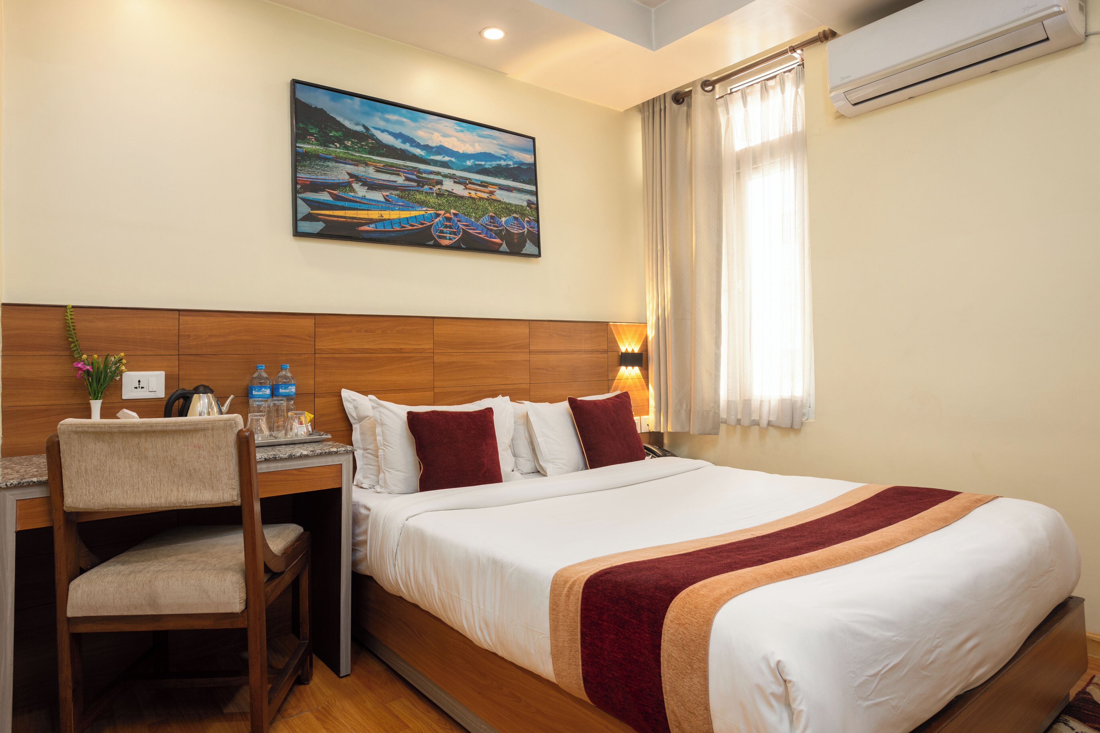 A hotel room with a bed, a desk, a chair, a painting on the wall, and an air conditioning unit.