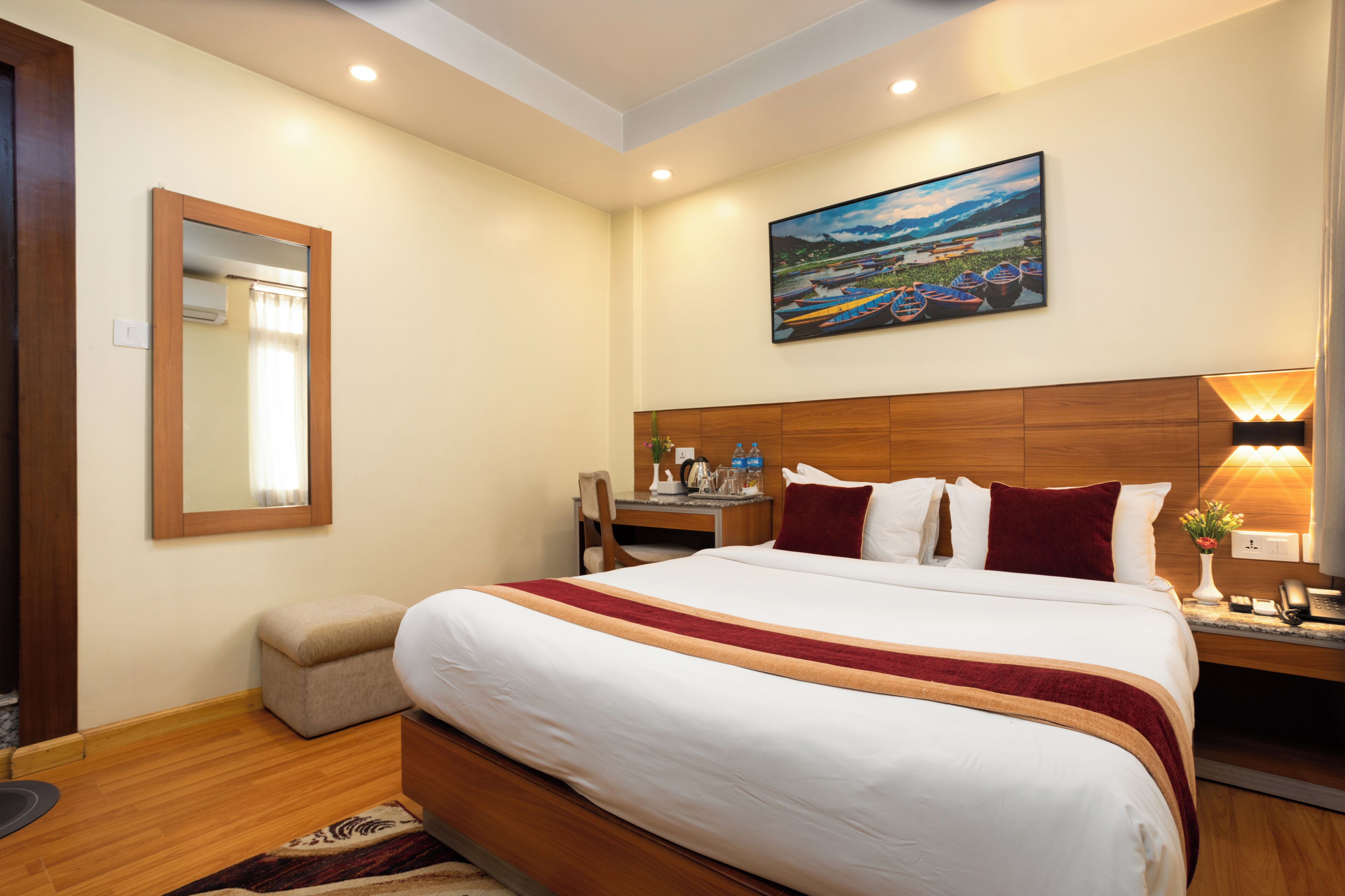 A hotel room with a large bed, a wooden headboard, a desk with a chair, a nightstand with a lamp, a painting on the wall, and a mirror.
