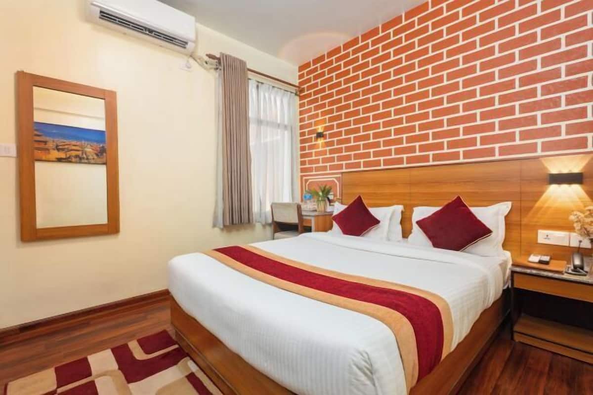 A hotel room with a brick wall, a bed with a red and white bedspread, a wooden headboard, a nightstand with a lamp, and a framed picture on the wall.